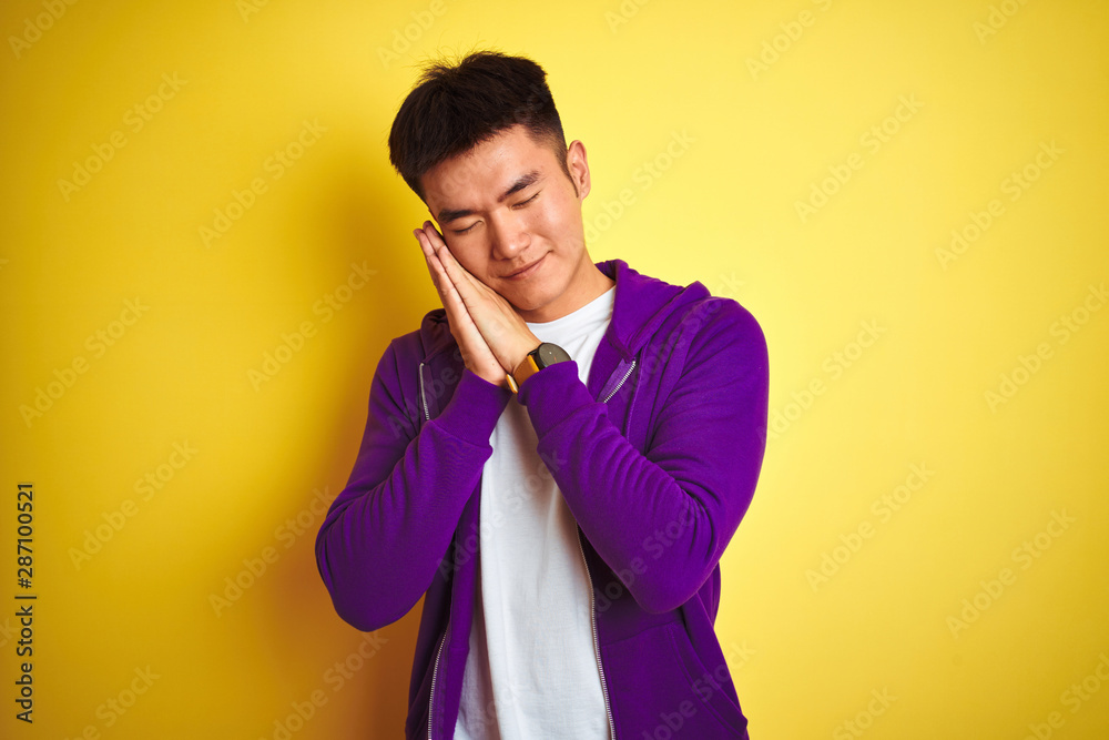 Young asian chinese man wearing purple sweatshirt standing over isolated yellow background sleeping tired dreaming and posing with hands together while smiling with closed eyes. Young asian chinese man wearing purple sweatshirt standing over isolated yellow background sleeping tired dreaming and posing with hands together while smiling with closed eyes.