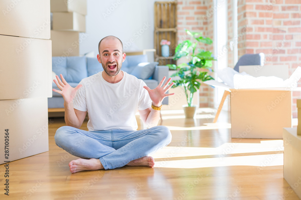 Young bald man sitting on the floor around cardboard boxes moving to a new home celebrating crazy and amazed for success with arms raised and open eyes screaming excited. Winner concept Young bald man sitting on the floor around cardboard boxes moving to a new home celebrating crazy and amazed for success with arms raised and open eyes screaming excited. Winner concept