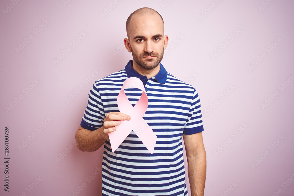 Young man holding pink brest cancer ribbon over isolated background with a confident expression on smart face thinking serious Young man holding pink brest cancer ribbon over isolated background with a confident expression on smart face thinking serious