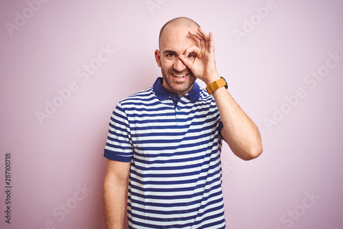 Young bald man with beard wearing casual striped blue t-shirt over pink isolated background doing ok gesture with hand smiling, eye looking through fingers with happy face.