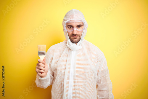 Young man wearing painter equipment and holding painting brush over isolated yellow background with a confident expression on smart face thinking serious