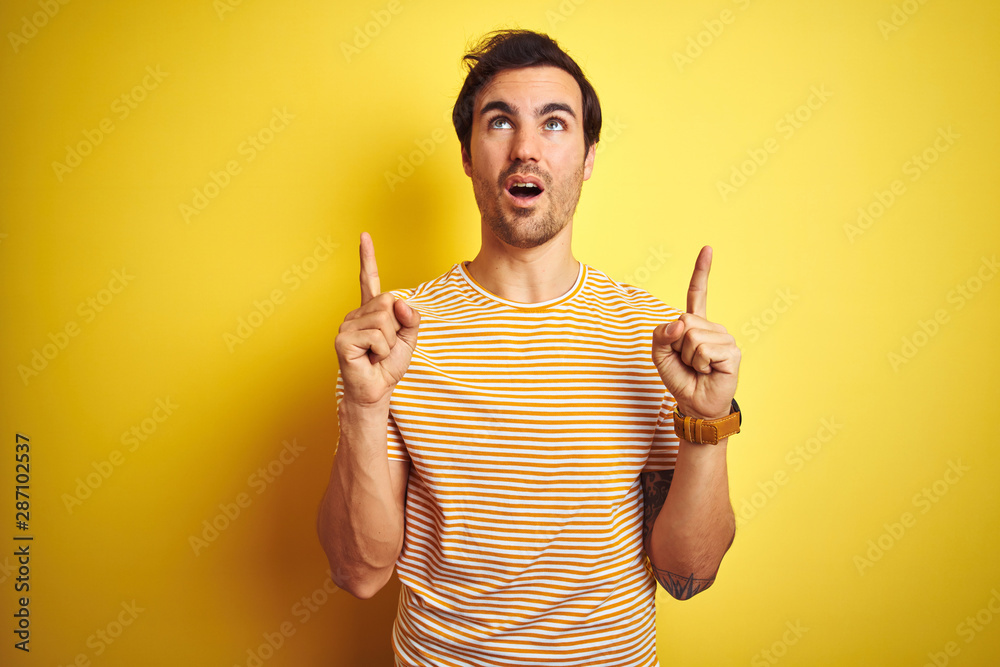 Young handsome man with tattoo wearing striped t-shirt over isolated yellow background amazed and surprised looking up and pointing with fingers and raised arms.