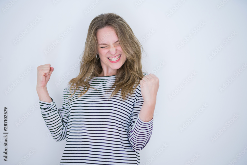 Beautiful blonde girl with blue eyes wearing striped sweater over white isolated background very happy and excited doing winner gesture with arms raised, smiling and screaming for success Beautiful blonde girl with blue eyes wearing striped sweater over white isolated background very happy and excited doing winner gesture with arms raised, smiling and screaming for success