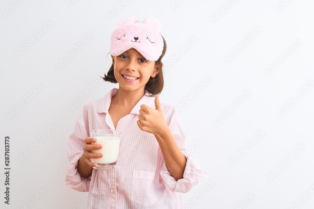 Child girl wearing sleep mask and pajama drinking glass of milk over isolated white background happy with big smile doing ok sign, thumb up with fingers, excellent sign Child girl wearing sleep mask and pajama drinking glass of milk over isolated white background happy with big smile doing ok sign, thumb up with fingers, excellent sign