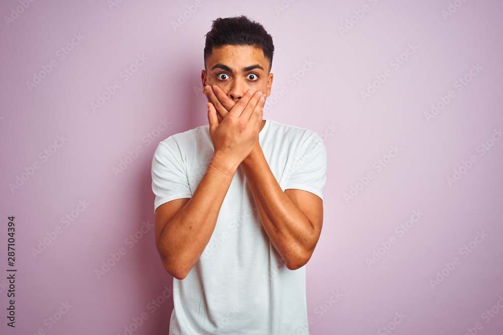Young brazilian man wearing t-shirt standing over isolated pink background shocked covering mouth with hands for mistake. Secret concept.