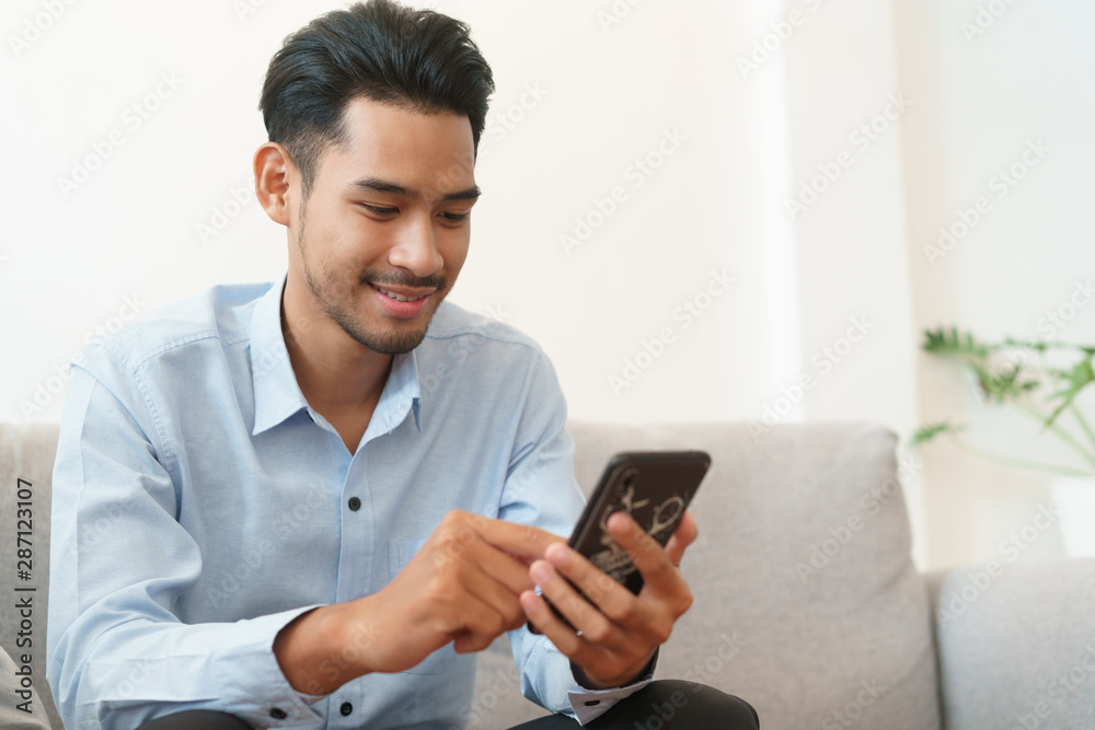 Portrait of young asian man handsome wearing blue shirt is reading using smartphone and sitting on sofa in living room at home Portrait of young asian man handsome wearing blue shirt is reading using smartphone and sitting on sofa in living room at home