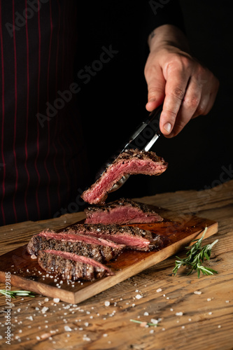 Slicing juicy beef steak by knife in chef hands closeup. Food cooking concept. Dark black background copy space.