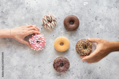 Hands holding fresh donuts on grey background with copy space. Assortment of various colourful donuts with different frosted, chocolate, pink glazed and sprinkles. Top view