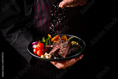 Grilled and sliced beef steak with grilled vegetables served on black plate on black background presentation in chef hands