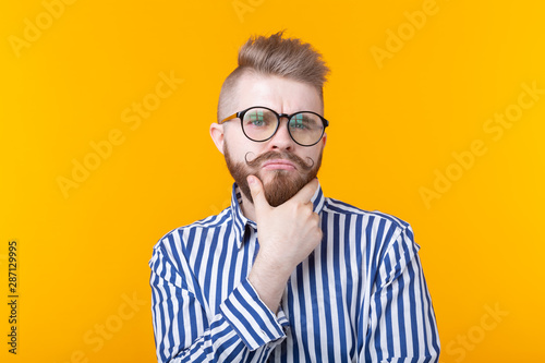 Cute intelligent young man with a beard and with glasses poses on a yellow background and thinks about something. The concept of a difficult choice. © satura_