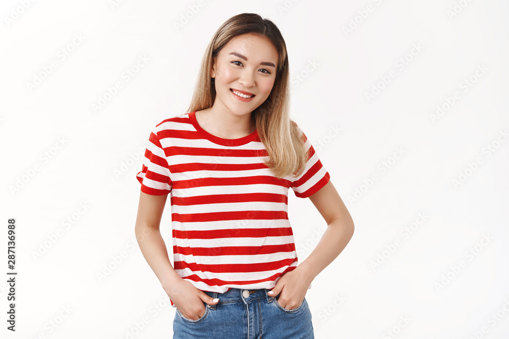 Fototapeta premium Friendly shy cute blond asian female hold hands jeans pockets smiling cheerful tilt head joyfully keep conversation talking campus neighbour standing white background modest asking out girl