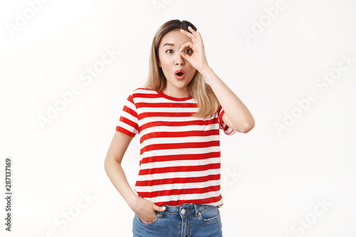 Wondered amused emotive blond asian woman drop jaw folding lips wow sound stare camera excited amazed show okay ok monocle gesture astonished look through fingers white background