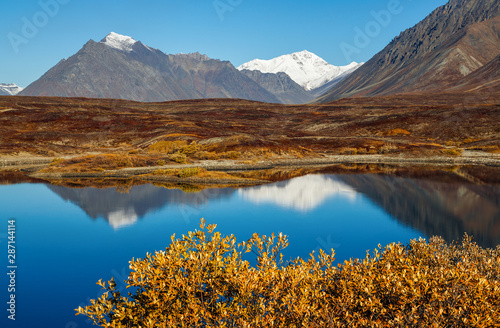 Picturesque autumn mountain landscape. Lake in a mountain valley. Snow-capped mountain peaks are reflected in the water. Travel and Hiking in the Arctic. Chukotka, Polar Siberia, Far East Russia.