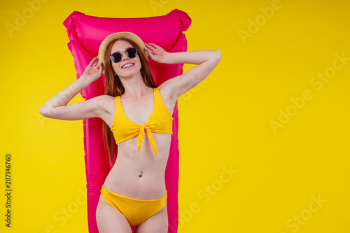 redhaired ginger female posing floating with inflatable mattress wearing swimmingsuit and hat,good looking in studio yellow background