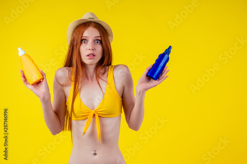amazed redhaired ginger woman holding two plastic bottle of suncream,facial expression "i don't know". copyspase studio yellow background