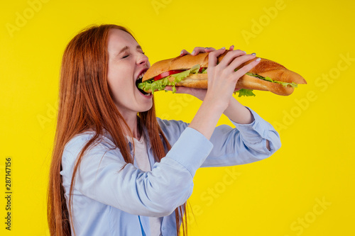 Joyful funny redhaired ginger girl with wild eyes suffering from anarexia with greedy ,eating large sandwic in studio yellow background. eats after 6 pm photo