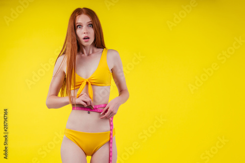 surprised and amazed redhaired ginger woman suffering eating disorders anorexia bulimia in white t-shirt and panties measuring waist with tape in studio yellow background