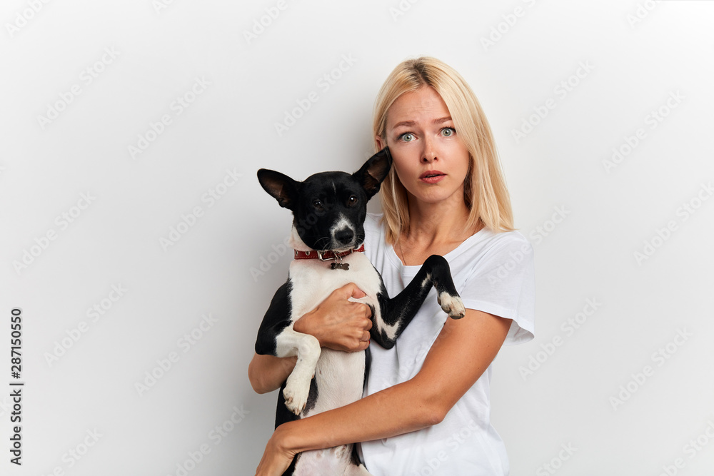 scared shocked woman carries lovely dog, dressed in white stylish T-shirt, has free time, isolated on white studio wall, close up portrait, facial expression, reaction