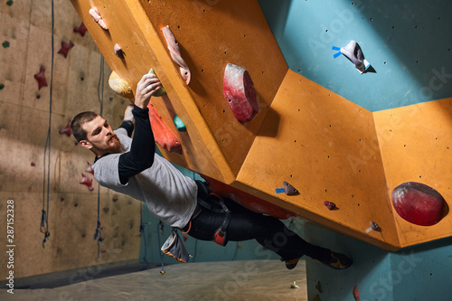 Side view of strong muscular physically challenged climber moving up on difficult steep rock, climbing on artificial wall in indoor bouldering gym, well-equipped, full length shot.