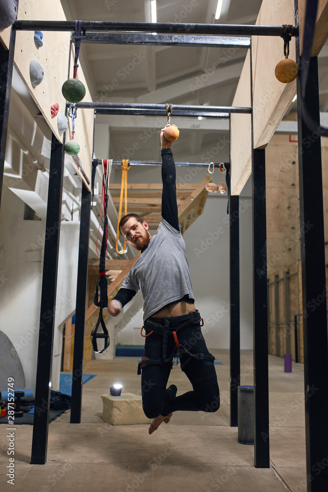 Cheerful handsome rock climber with physical disability training at chin up bar, hanging on one hand at suspended ball, doing special exercises for strength and endurance. Vertical shot, front view.