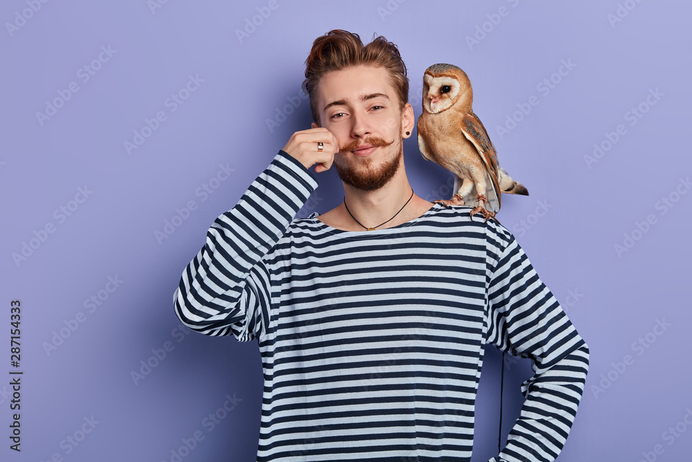 cheerful funny cunning guy in striped sweater taking money from tourist for taking a pictire with an owl, isolated blue background, studio shot, business concept - obrazy, fototapety, plakaty 