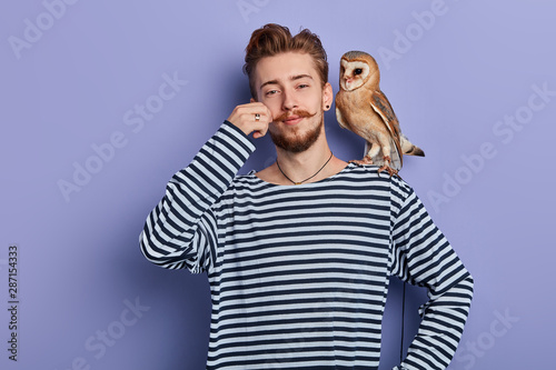 cheerful funny cunning guy in striped sweater taking money from tourist for taking a pictire with an owl, isolated blue background, studio shot, business concept photo