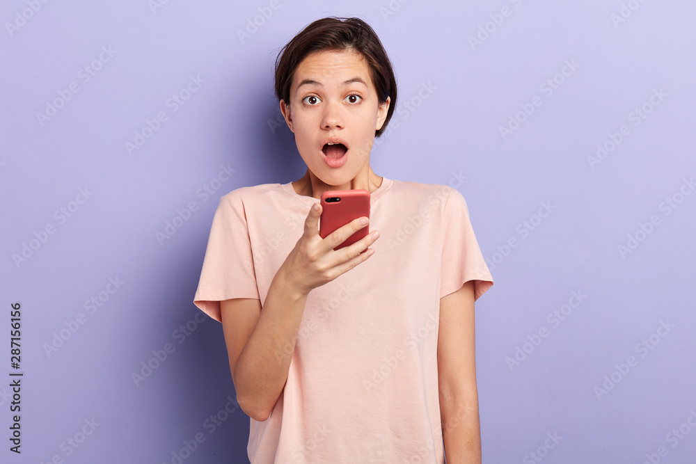 emotional girl holding smart phone and looking at the camera. woman cannot believe in news, information.isolated blue background, studio shot emotional girl holding smart phone and looking at the camera. woman cannot believe in news, information.isolated blue background, studio shot