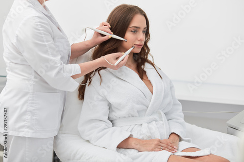 awesome good looking girl in white bathrobe receiving stimulating electric facial treatment from therapist, close up photo, wellness, wellbeing