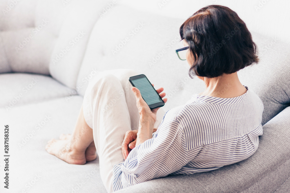 Middle-aged brunette woman with glasses on the gray sofa use smartphone. Middle-aged brunette woman with glasses on the gray sofa use smartphone.