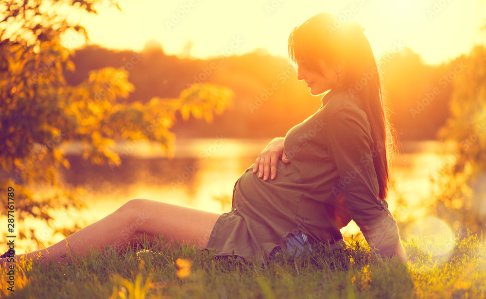 Pregnant woman sitting on green grass, looking on river on sunset, touching her belly. Enjoying healthy pregnancy