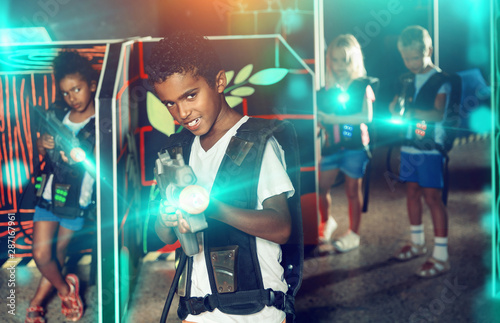 Emotional afro-american boy with laser pistol playing laser tag with friends on dark labyrinth