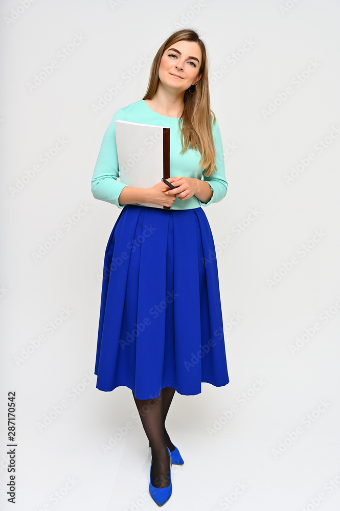 Full-length Vertical Portrait of a cute brunette middle-aged manager woman with beautiful curly hair in a business suit and blue skirt on a white background with a folder in her hands. Full-length Vertical Portrait of a cute brunette middle-aged manager woman with beautiful curly hair in a business suit and blue skirt on a white background with a folder in her hands.