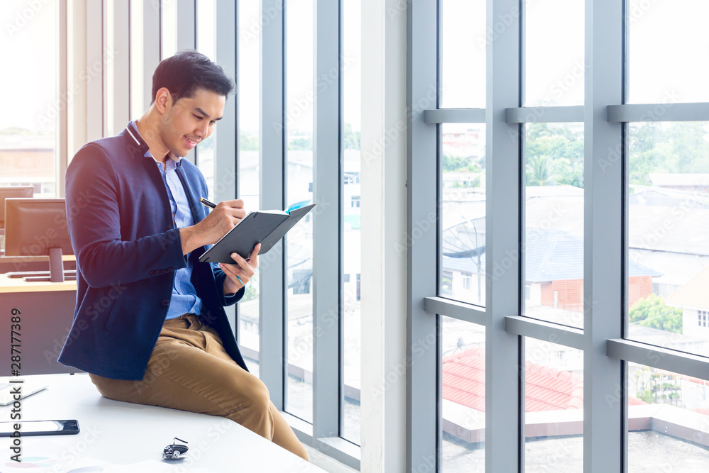 Fototapeta premium A businessman wearing a suit and a long-sleeve shirt inside and sitting smart poses in a modern office. In his hand have a pen and a notebook in action reading or writing something.
