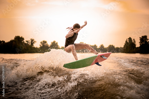 Skinny girl in black swimsuit jumping on the wakeboard on the river on the wave in the sunset