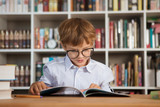 little boy wearing glasses reading book at table in library