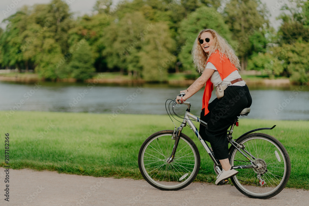 Outdoor shot of happy blonde curly woman wears sunglasses, dressed in casual active wears, rides on bicycle near lake and green trees in countryside, spends free time outside, enjoys favourite hobby Outdoor shot of happy blonde curly woman wears sunglasses, dressed in casual active wears, rides on bicycle near lake and green trees in countryside, spends free time outside, enjoys favourite hobby
