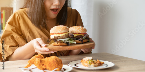 Hungry overweight woman holding hamburger on wooden plate, Fried chicken and Pizza on table .Concept of binge eating disorder (BED). photo