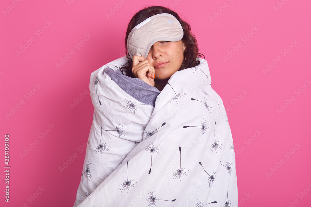Close up portrait of brunette woman peeping from sleeping mask, does not want to wake up, keeps eyes closed, wearing white blanket, isolated over pink studio wall. People, lifestyle, morning concept. Close up portrait of brunette woman peeping from sleeping mask, does not want to wake up, keeps eyes closed, wearing white blanket, isolated over pink studio wall. People, lifestyle, morning concept.