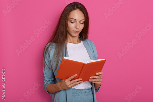 Close up portrait of young women reading something in notebook isolated over rosy background, looks concentrated, looking down, holding textbook organizer in hand, prepares to report. Studying concet