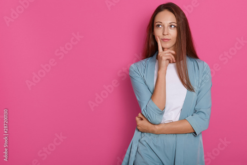 Studio shot of young female thinking and pondering over something with her finger on her chin, looking aside with pensive facial expression, wearing elegant outfit, isolated on pink background.