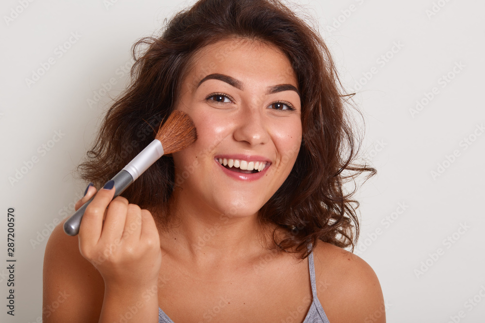 Image of Caucasian woman applying foundation powder with brush, model posing isolated over white background, stands smiling, preparing for dating, wearing gray t shirt. Makeup and beauty concept. Image of Caucasian woman applying foundation powder with brush, model posing isolated over white background, stands smiling, preparing for dating, wearing gray t shirt. Makeup and beauty concept.