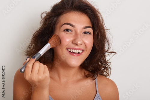 Image of Caucasian woman applying foundation powder with brush, model posing isolated over white background, stands smiling, preparing for dating, wearing gray t shirt. Makeup and beauty concept.