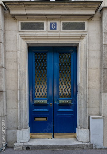 Vintage entrance and door windows with matte glass protected by rhombus patterned lattice. Bright blue painted antique door of old stone building in Paris France. Exterior of house in Europe © Ninel