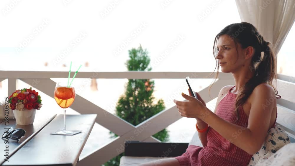 Beautiful girl with smartphone chilling in cafe. Inspired young woman in a red dress enjoying cocktail in restaurant on the seashore in summer weekend. Beautiful girl with smartphone chilling in cafe. Inspired young woman in a red dress enjoying cocktail in restaurant on the seashore in summer weekend.