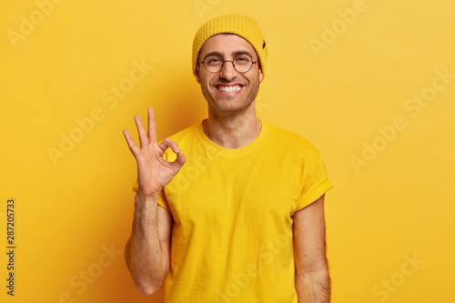 Portrait of handsome young man makes okay gesture, demonstrates agreement, likes idea, smiles happily, wears optical glasses, yellow hat and t shirt, models indoor. Its fine, thank you. Hand sign photo