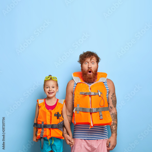 Surprised bearded dad with ginger beard, holds hand of little girl, wear protective lifejackets, ready for sea voyage, enjoy summer rest, stand against blue backgrond with copy space upwards
