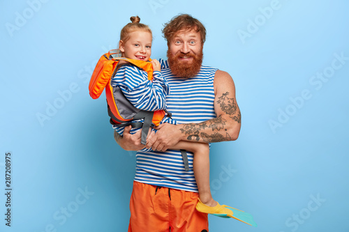 Cheerful young man poses with little ginger girl who wears orange lifevest, rubber fins, happy to spend summer holiday with father, enjoys swimming, isolated over blue background, giggle positively