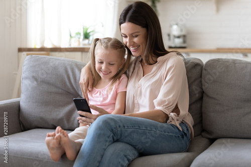 Positive mother embraces daughter sitting on couch using smartphone
