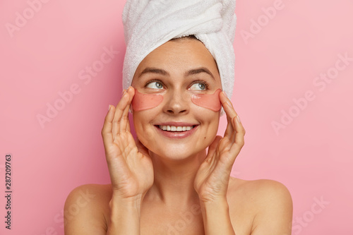 Positive pleasant looking woman cares about eyes skin beauty, appiles moisturizing collagen patches on face, looks aside, stands topless, has beauty treatments after taking bath, removes wrinkles