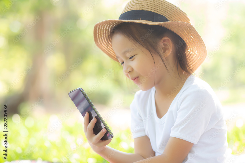 Beautiful asian child smiling using smart mobile phone in the garden, kid have passion playing game digital on smartphone on internet network in the park with happy, lifestyle and technology concept.